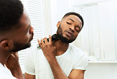 Young black man looking at mirror and shaving beard with trimmer