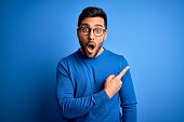 Young handsome man with beard wearing casual sweater and glasses over blue background Surprised pointing with finger to the side, open mouth amazed expression.