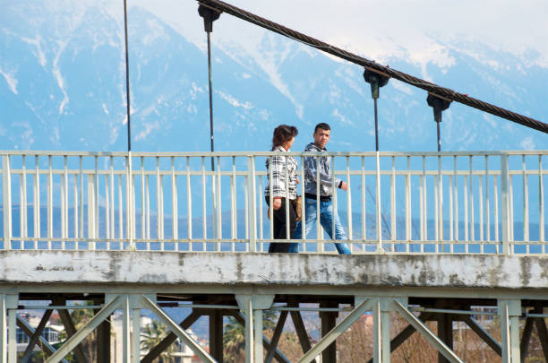 Young man and woman walking across suspension bridge before the snowcapped Tomorr mountain range.