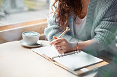 Young woman in blue warm sweater sitting near the big window of coffee shop and writing notes