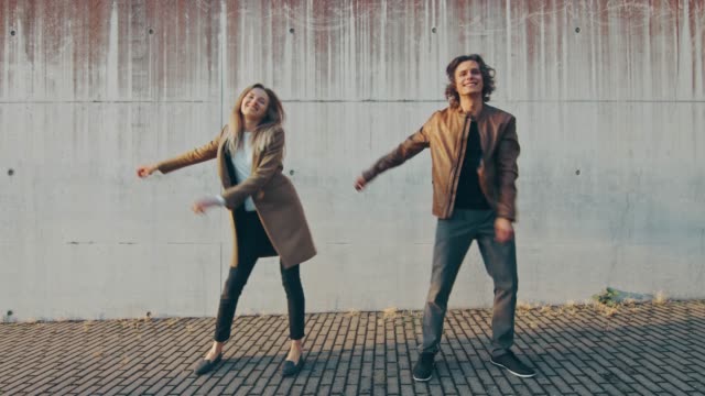 Cheerful Girl and Happy Young Man with Long Hair are Actively Dancing Meme Moves on a Street next to an Urban Concrete Wall. They Wear Brown Leather Jacket and Coat. Sunny Day.