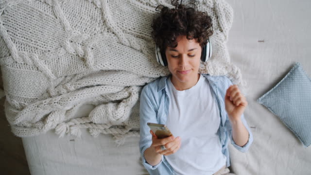 Slow motion of happy student in headphones using smartphone lying on bed at home