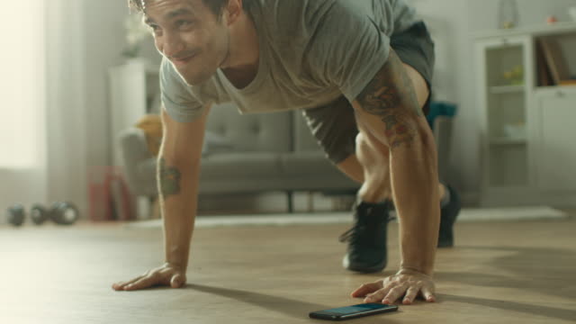 Slow Motion shot of a Muscular Fit Man in T-shirt and Shorts is Doing Mountain Climbers While Using a Stopwatch on His Phone. He is Training at Home in His Spacious and Bright Living Room with Minimalistic Interior.