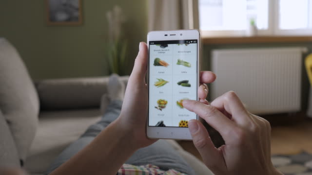 Woman Orders Food Home In An Online Store Using a Smartphone. Female Selects Vegetables in Grocery Online Store. Woman at Home Lying on Couch in Living Room Using Smartphone Buys in Internet Shop.