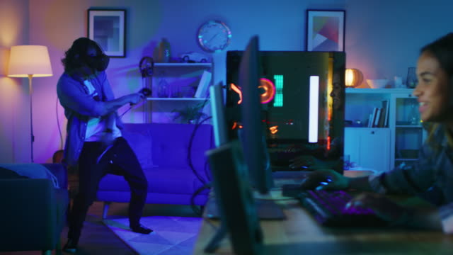 Young Man is Energetically Playing a Virtual Reality Video Game While Wearing a Headset. His Pretty Girl Friend Actively Plays an Online Shooter on a Computer in the Same Room with Neon Lights.